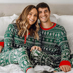 Couple wearing matching green and red Christmas pajamas sitting on a bed.